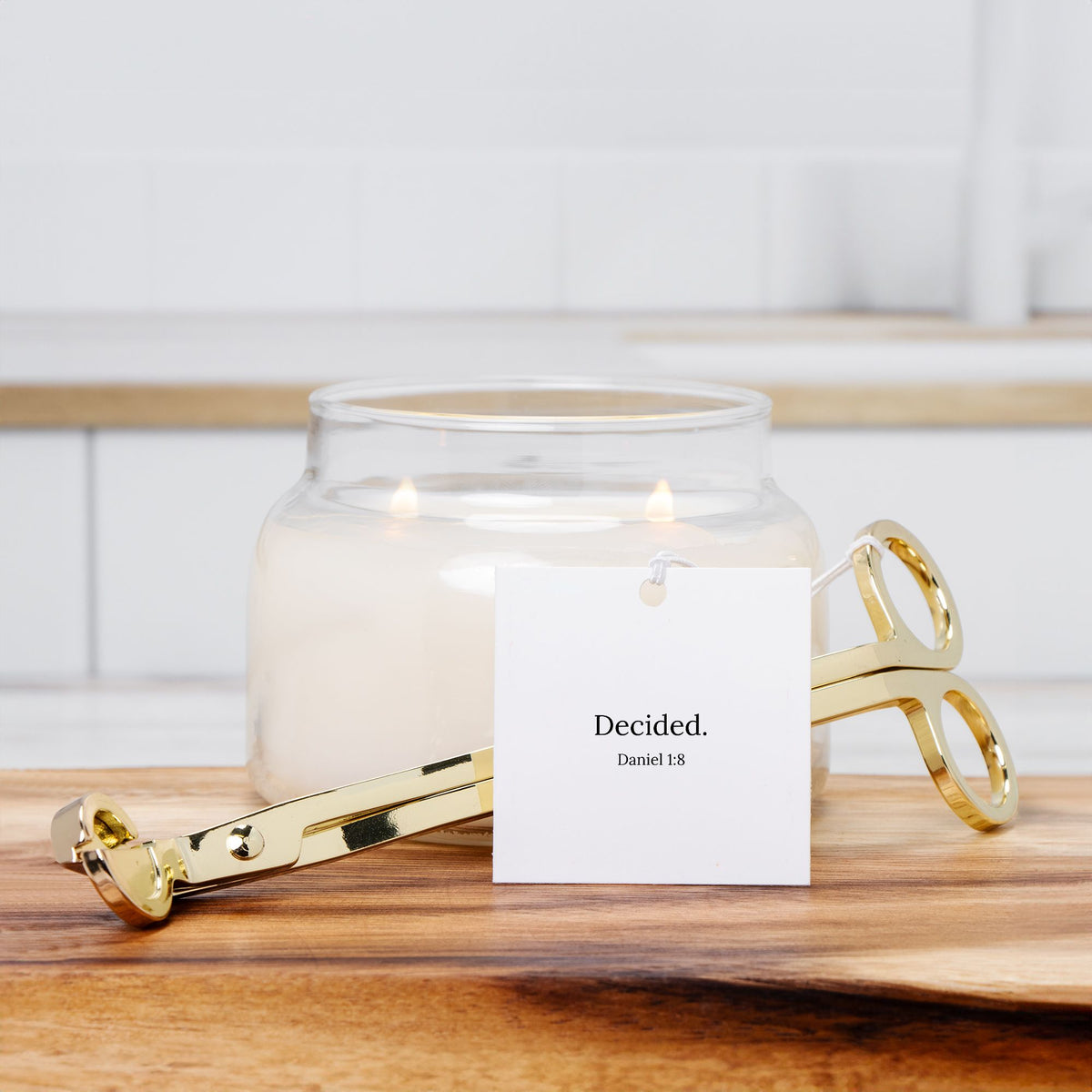 Gold wick trimmer with a DECIDED. tag beside a lit white jar candle